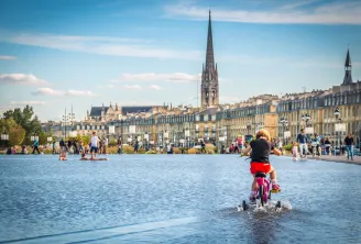 Enfant à vélo dans le miroir d'eau de Bordeaux avec la place de la Bourse et ses façades classiques en arrière-plan sous un ciel bleu dégagé.
