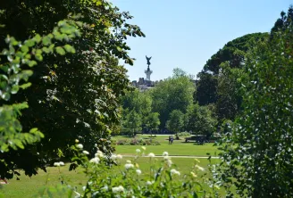 Vue à travers les arbres et la verdure d'un parc bordelais avec le Monument aux Girondins visible en arrière-plan, entouré de pelouses vertes et de visiteurs