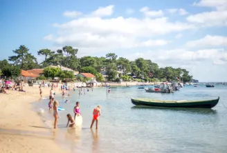 Plage animée avec baigneurs, bateaux de pêche colorés amarrés et maisons côtières sous les pins, par une belle journée ensoleillée