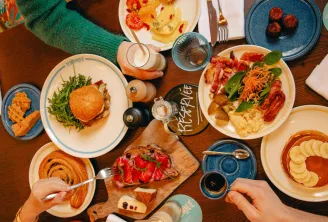 Vue aérienne d'une table de brunch à Bordeaux avec plusieurs assiettes colorées contenant un burger, des œufs, des fruits rouges, des croissants et des accompagnements variés, avec des mains de convives.