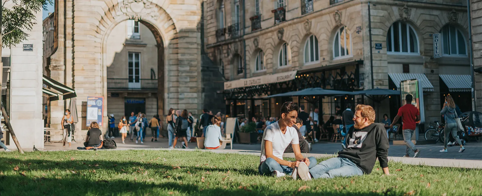 Deux jeunes hommes assis sur l'herbe conversent dans une place publique bordelaise, avec une architecture classique et une arche de pierre en arrière-plan, des passants et des commerces visibles.