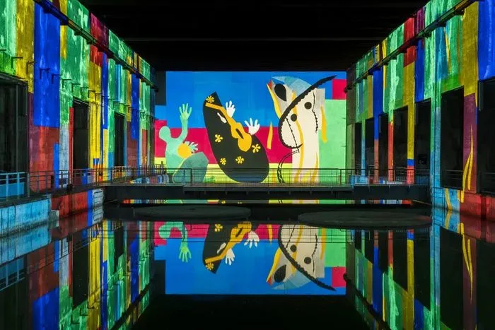 Installation immersive aux Bassins des Lumières de Bordeaux présentant une œuvre colorée inspirée de Matisse, avec des figures abstraites aux tons vifs (bleu, rouge, jaune, vert) projetées sur les murs et reflétées dans l'eau.