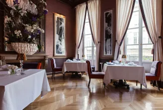 Élégante salle de restaurant gastronomique avec tables rondes dressées de nappes blanches, chaises bordeaux, grandes fenêtres avec rideaux crème, murs rose poudré, parquet en chevrons, et décoration florale raffinée.